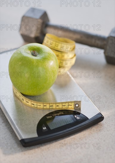 Green apple on weight scale, tape measure and exercise weight in background.