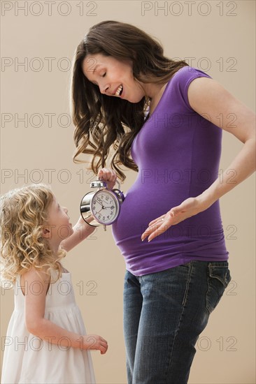 Girl (2-3) holding alarm clock next pregnant mother belly. Photo : Mike Kemp