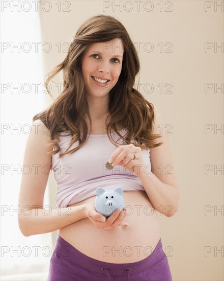 Young pregnant woman putting coin into blue piggybank. Photo : Mike Kemp