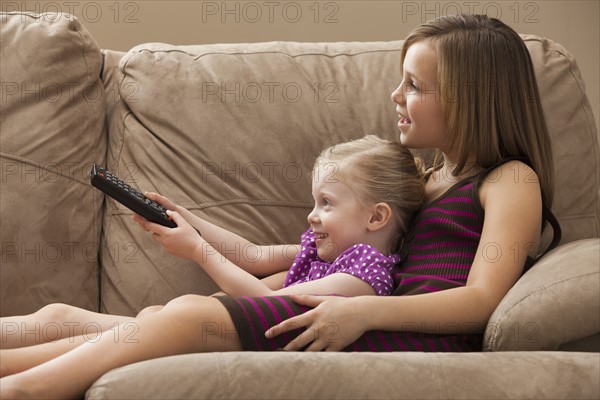 Girl (10-11) with sister (2-3) sitting on sofa, watching TV. Photo : Mike Kemp