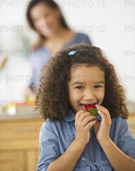 Smiling girl (6-7) eating strawberries with defocused woman in background.