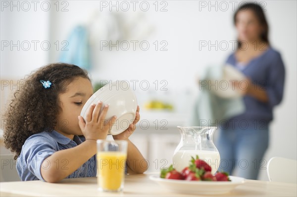 Girl (6-7) eating breakfast with defocused mother in background.