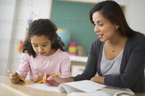 Girl (6-7) with female teacher sitting at desk in classroom.