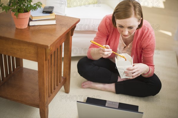 Woman sitting on floor, using laptop and eating take out food.