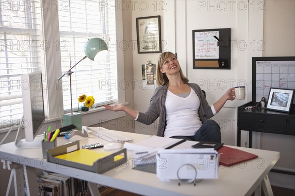 Woman sitting in office room, holding cup of tea, with arms stretched. Photo : DreamPictures