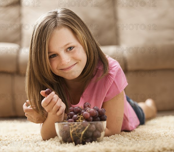Portrait of girl (10-11) lying on rug, eating red grapes. Photo : Mike Kemp