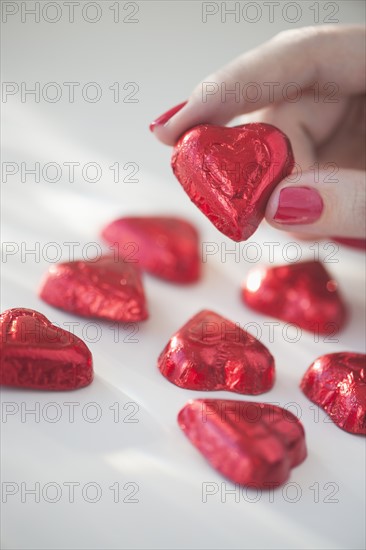 Close up of woman's fingers with red nail polish holding chocolate in shape of heart in red wrapping.