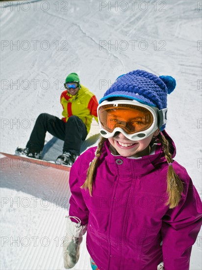 Daughter (10-11) posing with father sitting on ground with snowboard in background . Photo : db2stock