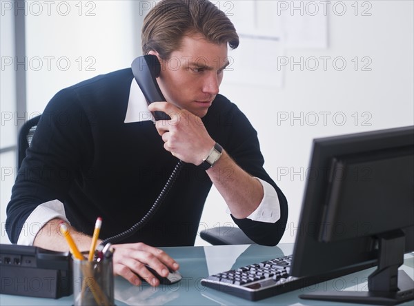 Man working on computer and talking on the phone at office. Photo : Daniel Grill