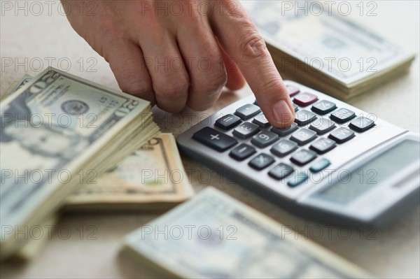 Close-up of man's hand using calculator and stacks of dollar bills.