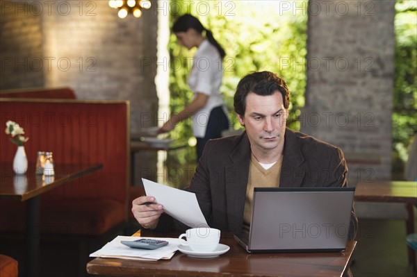 Owner working with laptop in restaurant with waitress in background.