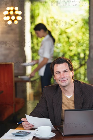 Owner working with laptop in restaurant with waitress in background.