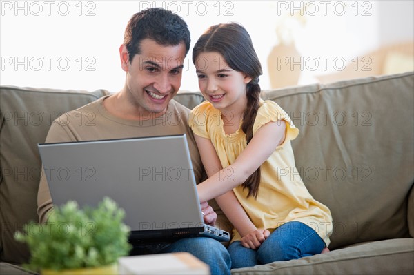 Daughter (8-9) and father sitting on sofa and using laptop .