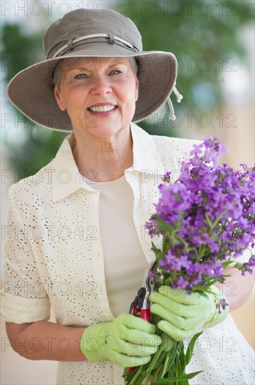 Portrait of smiling senior woman wearing gardening gloves, holding bunch of purple flowers.