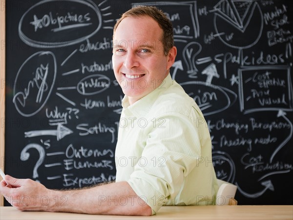 Businessman sitting at table against blackboard. Photo : Jamie Grill