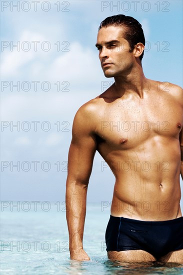 Sexy young man in swim trunks standing in the sea. Photo : momentimages