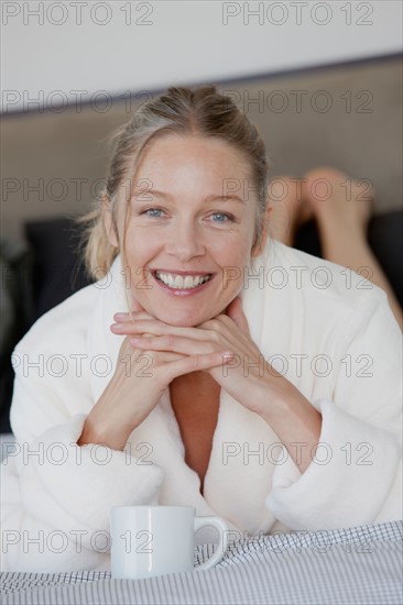 Portrait of mature woman with coffee cup lying on bed. Photo : Rob Lewine