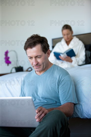 Mature man with laptop in bedroom and woman reading book in bed. Photo: Rob Lewine