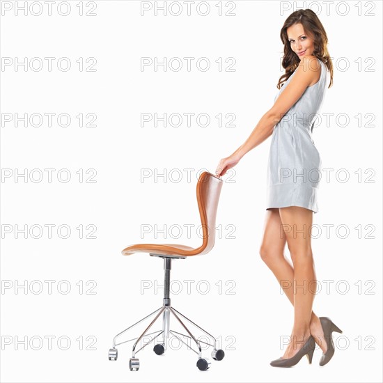 Studio shot of young elegant woman standing with hands on chair and smiling. Photo : momentimages