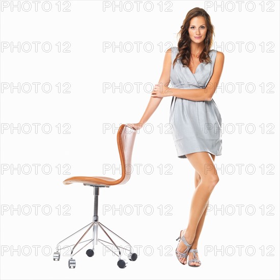 Studio shot of young elegant woman standing with hand on chair and smiling. Photo : momentimages