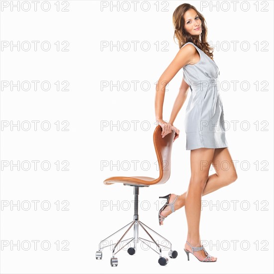 Studio shot of young elegant woman standing with hands on chair and smiling. Photo : momentimages