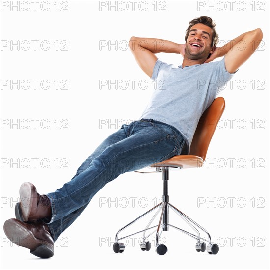 Portrait of young man relaxing on chair with hands behind head and smiling against white background. Photo : momentimages