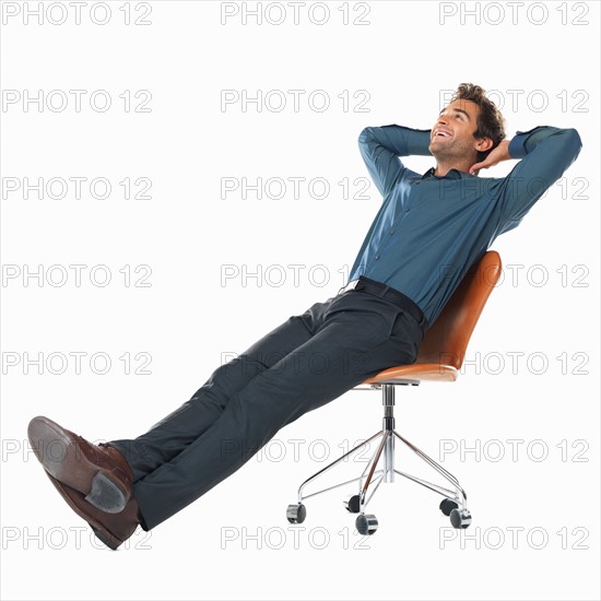 Studio shot of young man relaxing on chair with hands behind head. Photo: momentimages