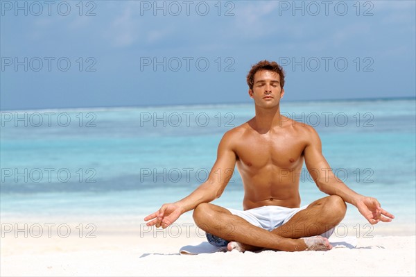 Handsome young man meditating in a lotus position on the beach. Photo: momentimages