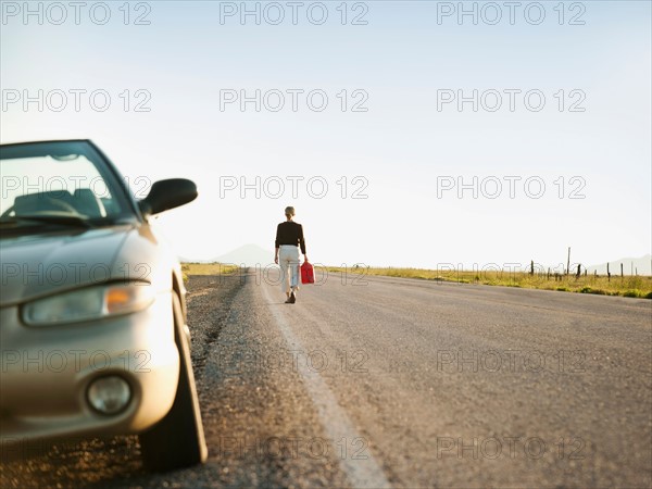 Woman carrying canister walking along empty road, her car parked on roadside.