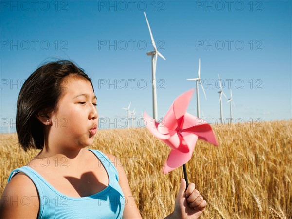 USA, Oregon, Wasco, Girl (10-11) holding blowing at fan in wheat field with wind turbines in background.