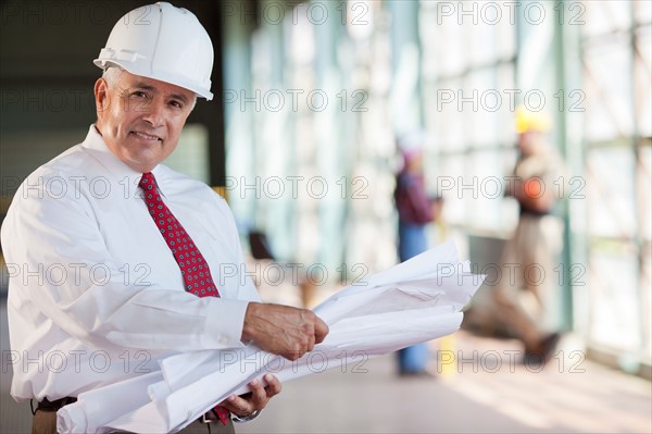 Portrait of senior man wearing tie and hardhat, holding blueprint. Photo: db2stock