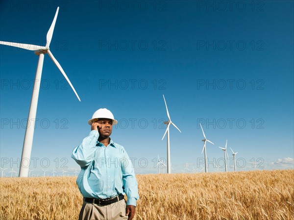 USA, Oregon, Wasco, Engineer standing in wheat field in front of wind turbines, using mobile phone.