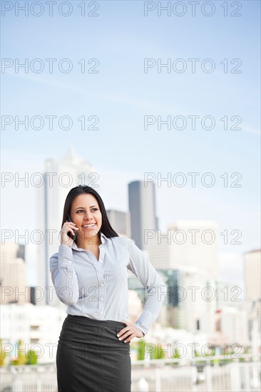 USA, Washington, Seattle, Young businesswoman talking on phone with city skyline behind. Photo : Take A Pix Media