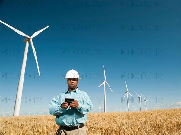 USA, Oregon, Wasco, Engineer standing in wheat field in front of wind turbines, using mobile phone.