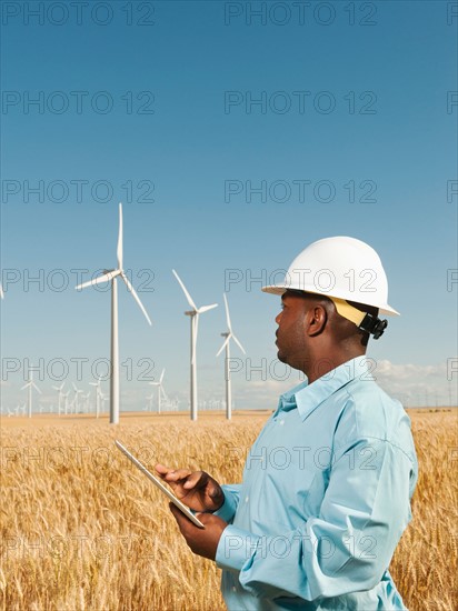 USA, Oregon, Wasco, Engineer standing in wheat field in front of wind turbines, using digital tablet.
