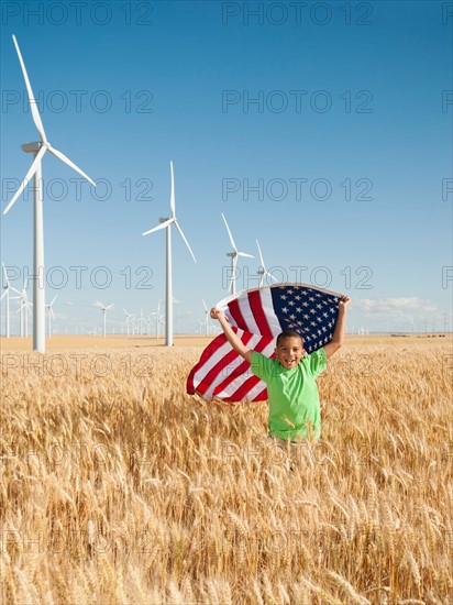 USA, Oregon, Wasco, Boy (8-9) flying american flag in wheat field with wind turbines in background.