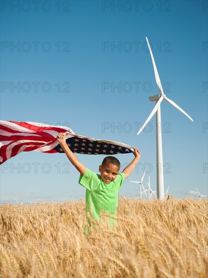 USA, Oregon, Wasco, Boy (8-9) flying american flag in wheat field with wind turbines in background.