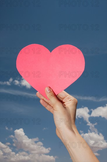 Woman's hand holding pink heart shape against sky. Photo : Winslow Productions