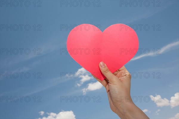 Woman's hand holding pink heart shape against sky. Photo : Winslow Productions