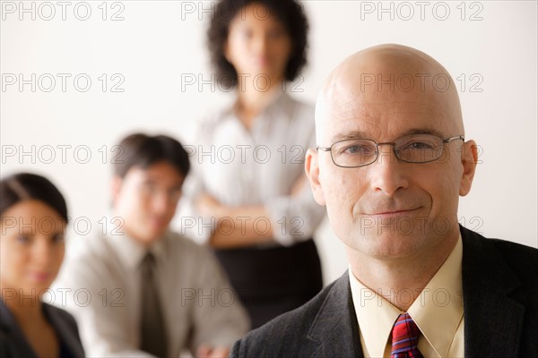 Mature businessman looking at camera, business team in background. Photo : Rob Lewine