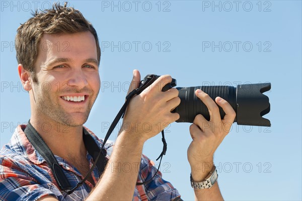 Portrait of man holding camera against blue sky.