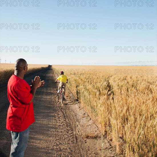 Father clapping as son (8-9) is cycling along dirt road in fields.