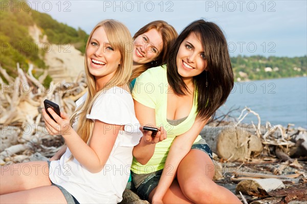 Three young women hanging out on beach, using cell phones. Photo: Take A Pix Media