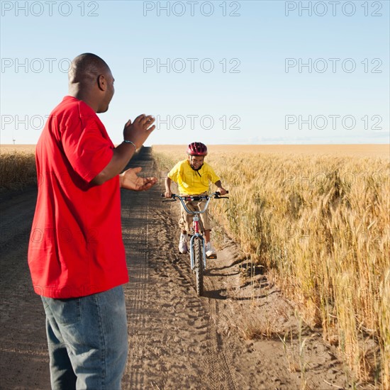 Father clapping as son (8-9) is cycling along dirt road in fields.