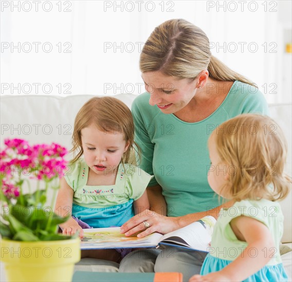 Mother with daughters (2-3) reading book on sofa.