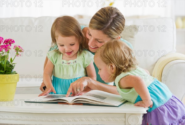 Mother with daughters (2-3) reading book in living room.