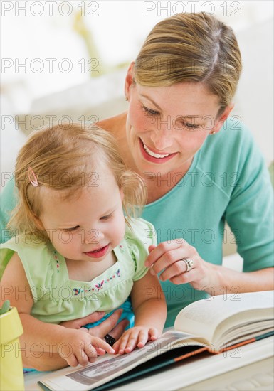 Mother with daughter (2-3) reading book in living room.
