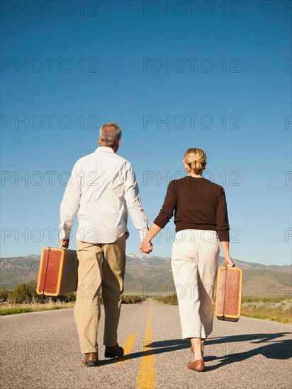 Mid adult couple walking along empty road carrying suitcases. Photo: Erik Isakson