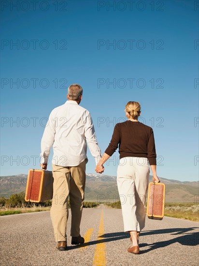 Mid adult couple walking along empty road carrying suitcases.