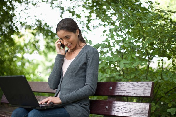 Young woman sitting on park bench using laptop and cell phone. Photo : Jan Scherders